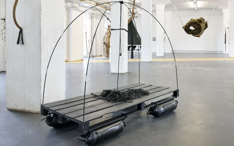 Zu sehen ist "Traveling Light", eine raumgreifende Installation, die aus einer rechteckigen palettenartigen Plattform aus schwarz-lackiertem Holz besteht, welches auf schwarzen Schwimmkörpern ruht und mit schwarzen Metall- oder Plastikbögen überspannt ist, die sich oben in der Mitte kreuzen. Auf der schwarzen Plattform liegt ein Bündel schwarzes Seil. Die Installation befindet sich in einem Ausstellungskontext in einer großen Halle mit dunkelgrauem Boden, weißen Wänden und mehreren weißen Stützpfeilern. Rechts im Hintergrund hängt ein massiver Baumstamm von der Decke, der innen hohl ist, und im Hintergrund wird der Raum von einer gelben Bahn durchquert, die über den Hallenboden verläuft.